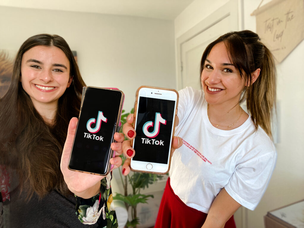 Die beiden TikTokerinnen halten ihre Smartphones in die Kamera. Darauf ist das TikTok-Logo zu sehen.
